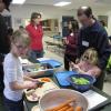 Several adults help children prepare carrots and celery in a kitchen area.
