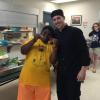 A student wearing a yellow t-shirt holds up two vegetables poses next to a man in a black chef uniform for a photo.