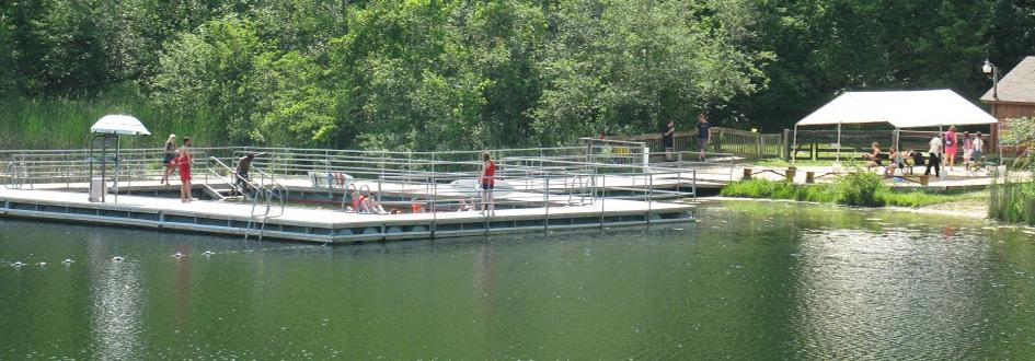 Several lifeguards stand on a large wooden dock lined with metal railings and a small pool in the center. Adults and children sit on picnic benches under a white tent on shore.